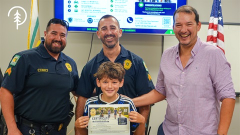 photo of two officers with a child holding a certificate of completion for the ebike safety class and his father/guardian next to him